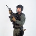 A male individual in full tactical law enforcement or military-style gear is posed against a plain white studio background. He is holding an assault-style rifle in a ready position and is equipped with extensive tactical equipment. The image appears to be a professional studio photograph, likely for promotional or documentation purposes.