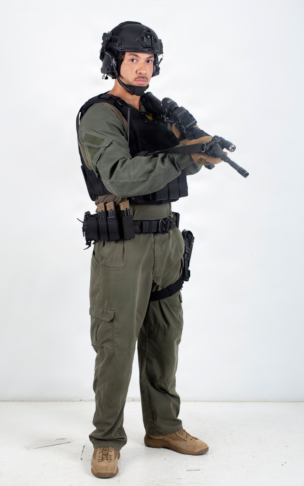 A male individual is posed against a plain white studio background wearing full tactical/law enforcement gear. He is holding a rifle with mounted accessories in a low-ready or patrol carry position. The image appears to be a professional equipment or personnel documentation photograph.