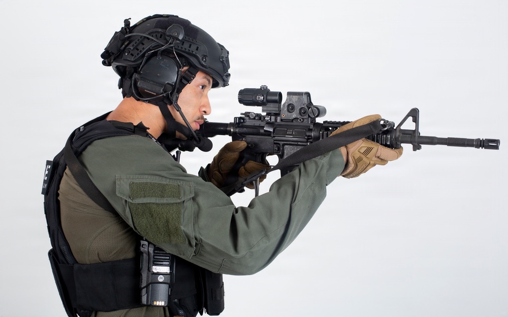 A tactical operator or law enforcement personnel is shown in a side-profile stance aiming an AR-15/M4-style rifle. The individual is equipped with full tactical gear including a ballistic helmet, body armor, and communications equipment. The image appears to be a professional studio or product photograph taken against a plain white background.