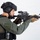 MCAS Marines pose for content 2026 A tactical operator or law enforcement personnel is shown in a side-profile stance aiming an AR-15/M4-style rifle. The individual is equipped with full tactical gear including a ballistic helmet, body armor, and communications equipment. The image appears to be a professional studio or product photograph taken against a plain white background.