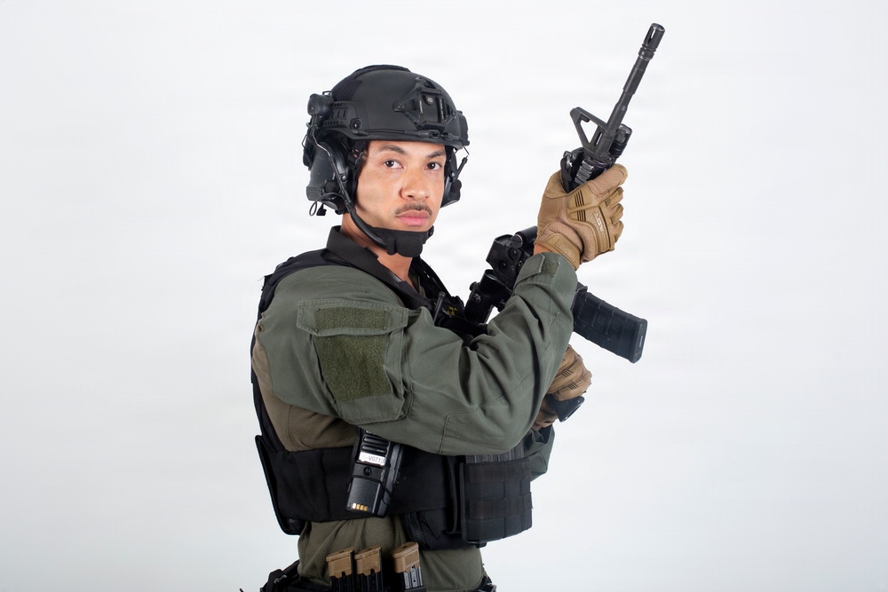 A person in full tactical/military gear is posed against a plain white studio background. They are holding a rifle in a raised, ready position while looking directly at the camera. The image appears to be a professional studio photograph, likely for commercial or editorial purposes.