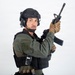 A person in full tactical/military gear is posed against a plain white studio background. They are holding a rifle in a raised, ready position while looking directly at the camera. The image appears to be a professional studio photograph, likely for commercial or editorial purposes.