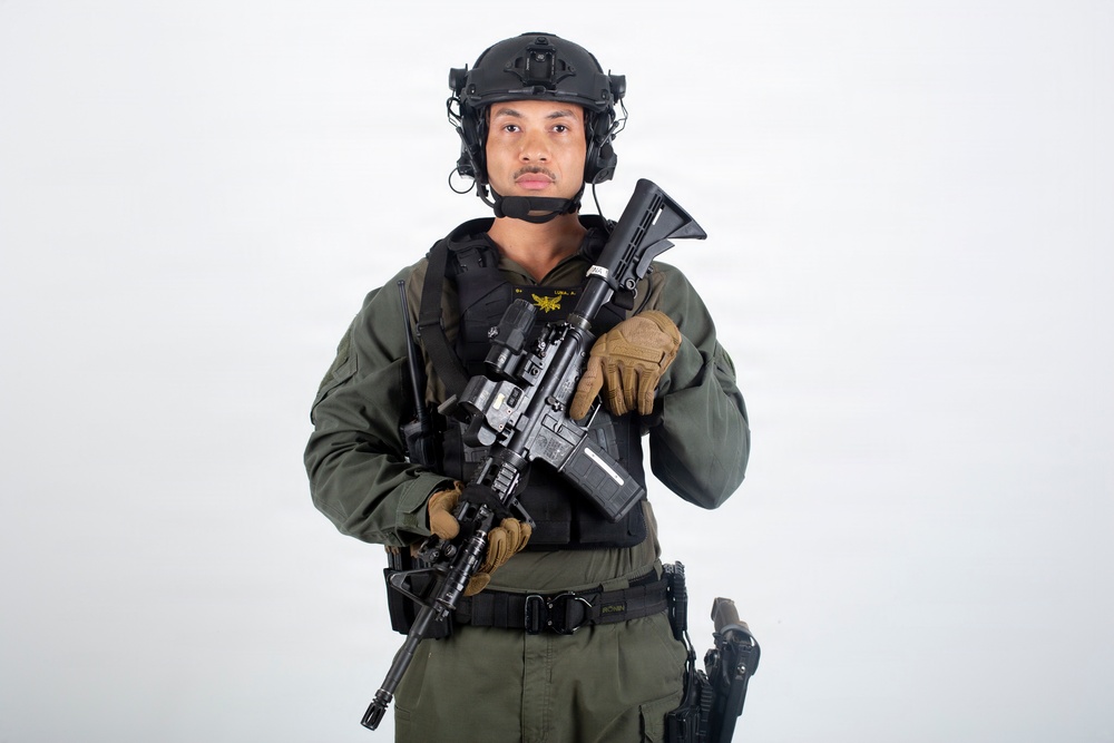 A male individual in full tactical/military gear is posed against a plain white background in what appears to be a professional studio photograph. He is wearing olive drab fatigues, a black tactical vest, and a black ballistic helmet with communications equipment. He is holding an assault rifle with accessories across his chest in a ready/display position.