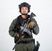 A male individual in full tactical/military gear is posed against a plain white background in what appears to be a professional studio photograph. He is wearing olive drab fatigues, a black tactical vest, and a black ballistic helmet with communications equipment. He is holding an assault rifle with accessories across his chest in a ready/display position.