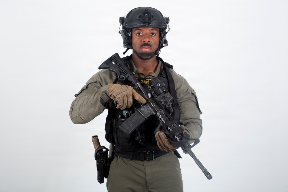 A male individual in full tactical/military gear is posed against a plain white background. He is equipped with a rifle, tactical vest, helmet with night-vision mount, and sidearm holster. The image appears to be a professional or studio-style portrait photograph.