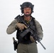 A male individual in full tactical/military gear is posed against a plain white background. He is equipped with a rifle, tactical vest, helmet with night-vision mount, and sidearm holster. The image appears to be a professional or studio-style portrait photograph.