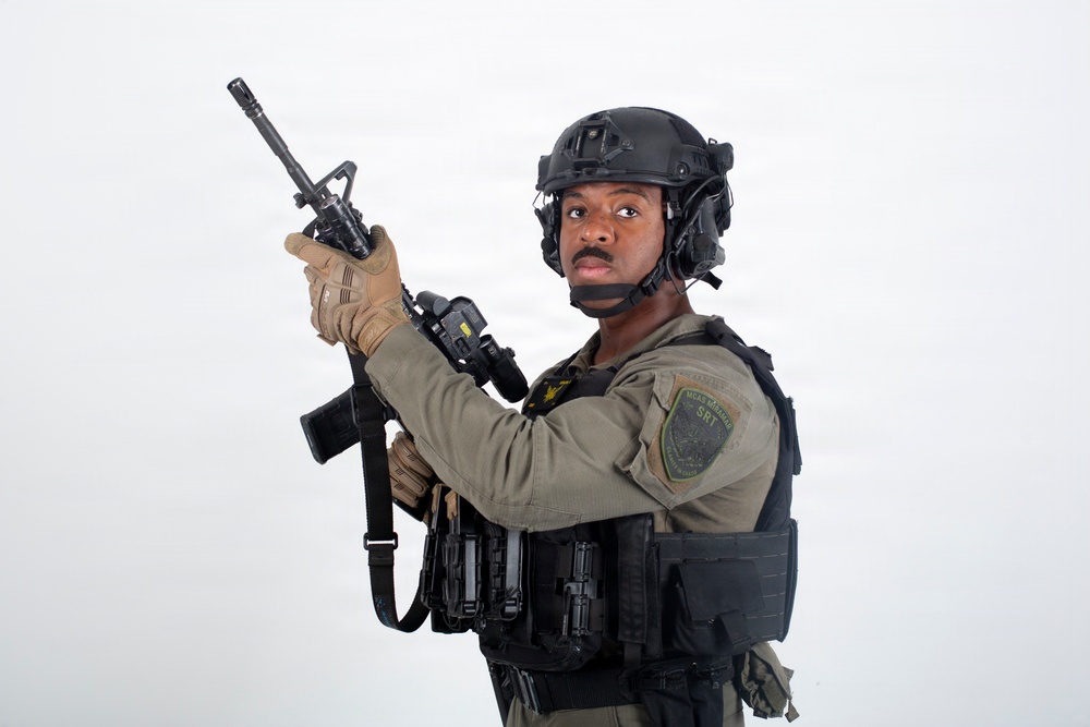 A uniformed tactical officer is posed against a white background holding a rifle in an upward position. The individual is wearing full tactical gear including a ballistic helmet, body armor, and communications equipment. A shoulder patch reading 'SRT' (Special Response Team) is visible on the uniform.