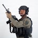 A uniformed tactical officer is posed against a white background holding a rifle in an upward position. The individual is wearing full tactical gear including a ballistic helmet, body armor, and communications equipment. A shoulder patch reading 'SRT' (Special Response Team) is visible on the uniform.