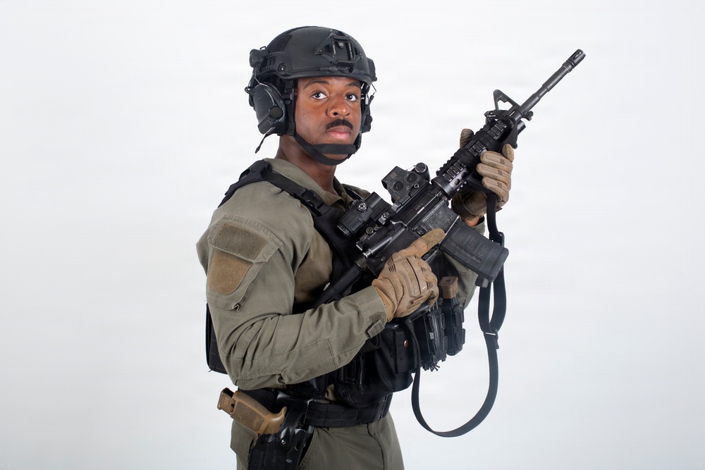 A soldier or tactical operator is posed against a plain white/light gray studio background in full combat gear. The subject is holding an assault rifle in a ready position while looking toward the camera. The image appears to be a professional studio photograph, likely for editorial, stock, or promotional purposes.