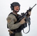 A soldier or tactical operator is posed against a plain white/light gray studio background in full combat gear. The subject is holding an assault rifle in a ready position while looking toward the camera. The image appears to be a professional studio photograph, likely for editorial, stock, or promotional purposes.