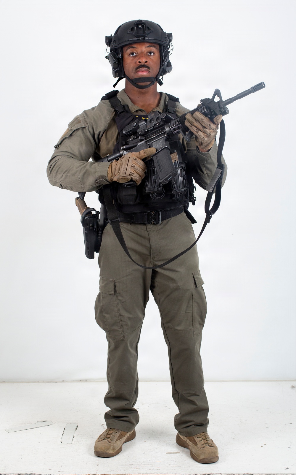 A young male individual is photographed against a plain white studio background in full tactical gear. He is wearing olive/ranger green uniform with black plate carrier vest, ballistic helmet with accessories, and is holding a short-barreled rifle with optics. The image appears to be a professional equipment or catalog-style photograph.