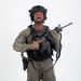 A young male individual is photographed against a plain white studio background in full tactical gear. He is wearing olive/ranger green uniform with black plate carrier vest, ballistic helmet with accessories, and is holding a short-barreled rifle with optics. The image appears to be a professional equipment or catalog-style photograph.