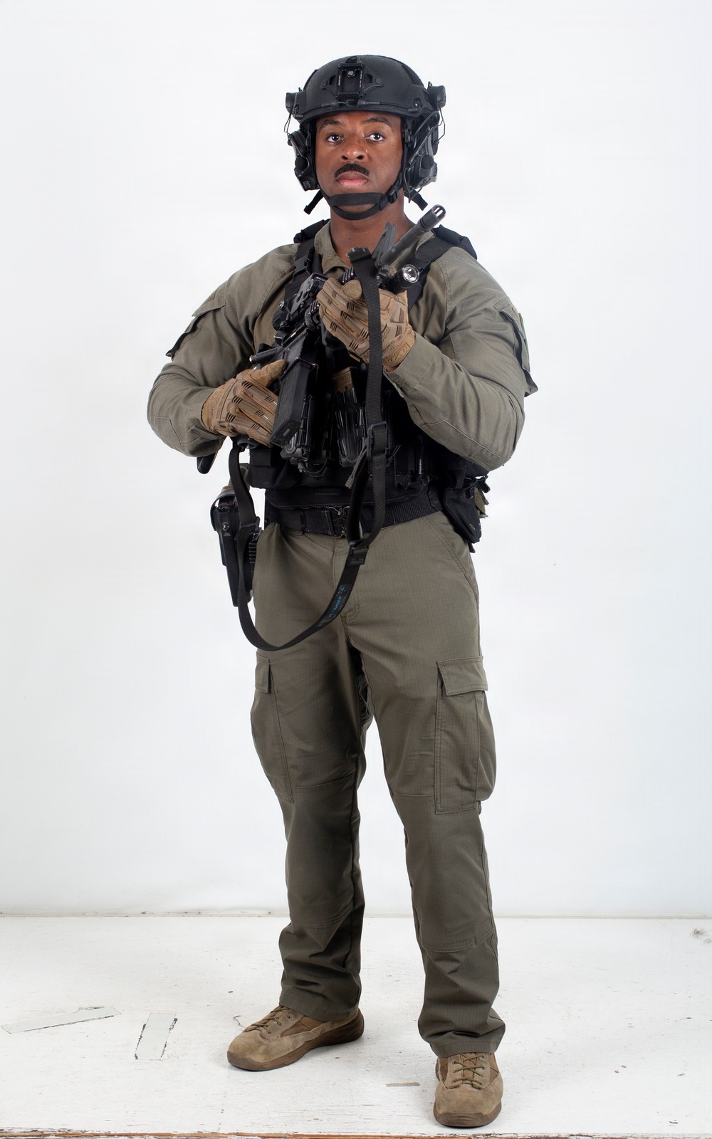 A male individual is photographed against a plain white studio background in full tactical gear. He is wearing olive/sage green combat clothing with a black tactical vest, black ballistic helmet, and is holding a firearm at chest level. The image appears to be a professional equipment or personnel documentation photograph.