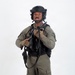 A male individual is photographed against a plain white studio background in full tactical gear. He is wearing olive/sage green combat clothing with a black tactical vest, black ballistic helmet, and is holding a firearm at chest level. The image appears to be a professional equipment or personnel documentation photograph.