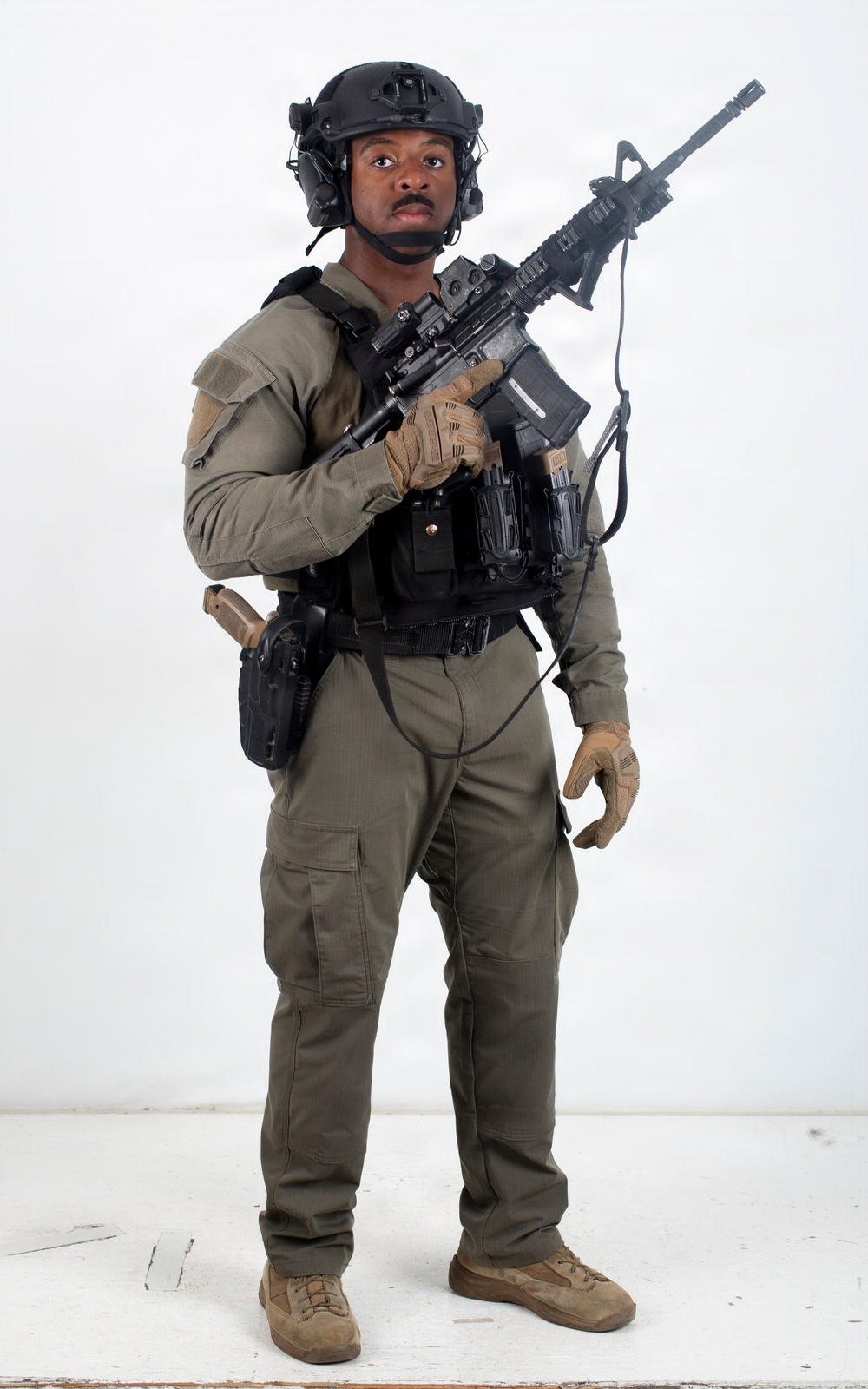 A male individual is posed against a plain white studio background wearing full tactical gear. He is holding an assault-style rifle in a low-ready or patrol carry position while equipped with a plate carrier, helmet, and sidearm. The image appears to be a professional studio photograph, likely for promotional, training, or catalog purposes.
