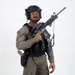 A male individual is posed against a plain white studio background wearing full tactical gear. He is holding an assault-style rifle in a low-ready or patrol carry position while equipped with a plate carrier, helmet, and sidearm. The image appears to be a professional studio photograph, likely for promotional, training, or catalog purposes.