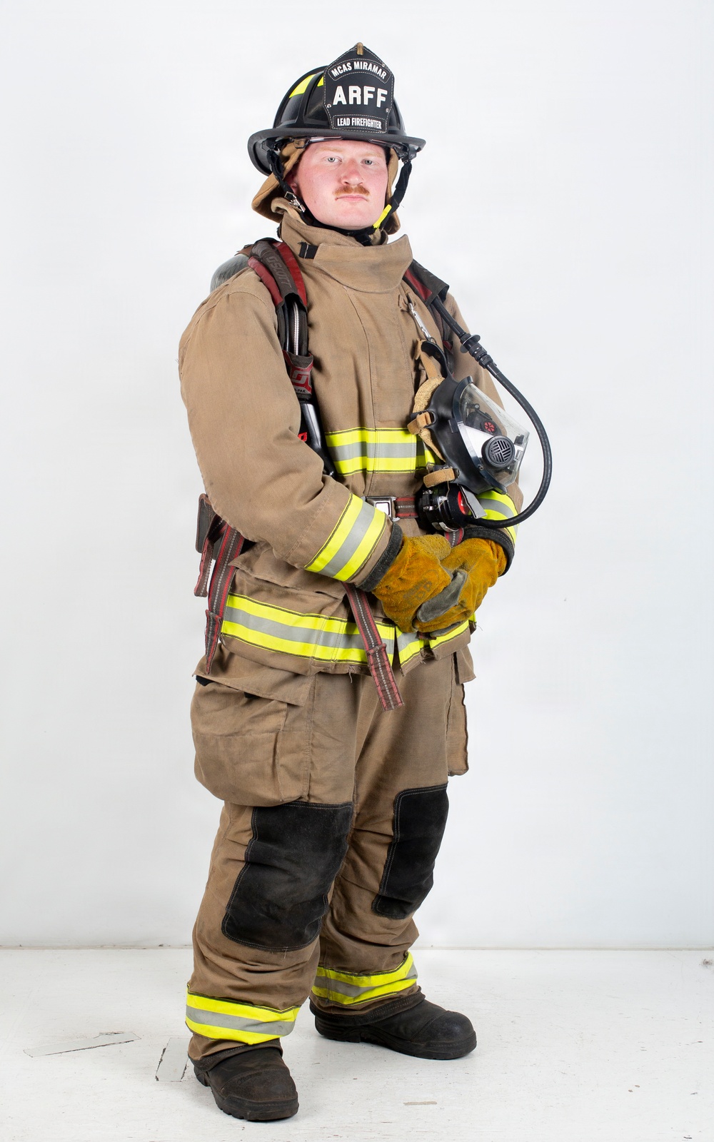 A firefighter in full turnout gear stands against a plain white background in a formal portrait-style photograph. The individual is wearing a black helmet labeled 'ARFF' and 'MCAS Miramar - Lead Firefighter,' indicating affiliation with Aircraft Rescue and Firefighting at Marine Corps Air Station Miramar. The subject holds an SCBA (Self-Contained Breathing Apparatus) face mask and is fully equipped with protective gear.