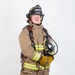 A firefighter in full turnout gear stands against a plain white background in a formal portrait-style photograph. The individual is wearing a black helmet labeled 'ARFF' and 'MCAS Miramar - Lead Firefighter,' indicating affiliation with Aircraft Rescue and Firefighting at Marine Corps Air Station Miramar. The subject holds an SCBA (Self-Contained Breathing Apparatus) face mask and is fully equipped with protective gear.