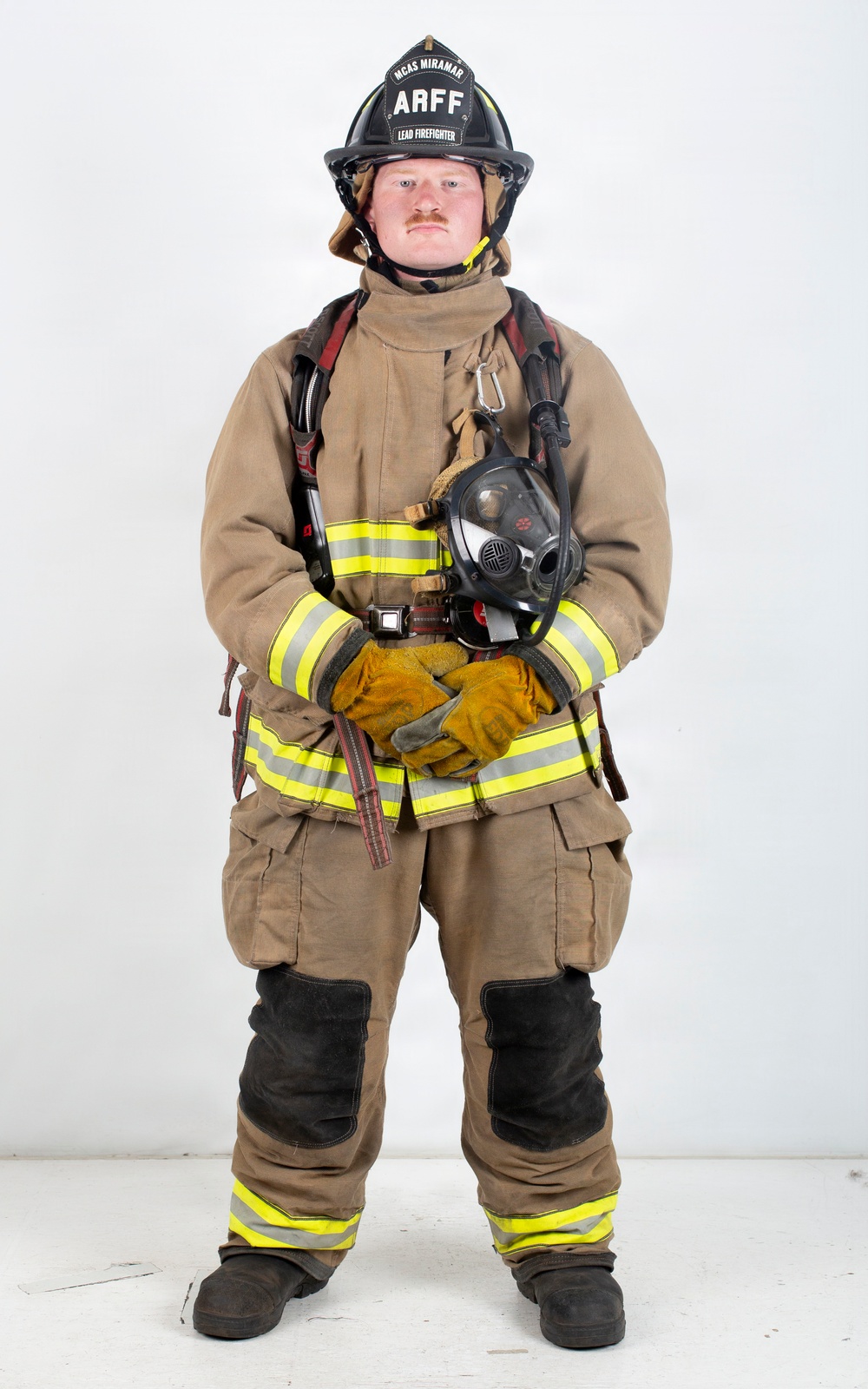 A firefighter in full personal protective equipment stands in a formal portrait pose against a plain white background. The helmet identifies the individual as a Lead Firefighter with ARFF (Aircraft Rescue and Firefighting) at MCAS Miramar. The subject is holding a breathing apparatus mask and wearing complete turnout gear.