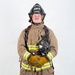 A firefighter in full personal protective equipment stands in a formal portrait pose against a plain white background. The helmet identifies the individual as a Lead Firefighter with ARFF (Aircraft Rescue and Firefighting) at MCAS Miramar. The subject is holding a breathing apparatus mask and wearing complete turnout gear.