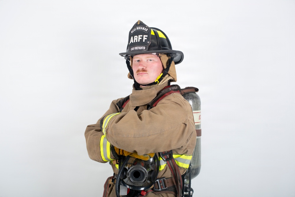A firefighter in full turnout gear poses with arms crossed against a plain white background. The subject wears a black helmet labeled 'ARFF' and 'MCAS Miramar' with 'Lead Firefighter' designation, indicating affiliation with Aircraft Rescue and Firefighting at Marine Corps Air Station Miramar. The professional portrait-style photo captures the firefighter in complete personal protective equipment including an SCBA tank.
