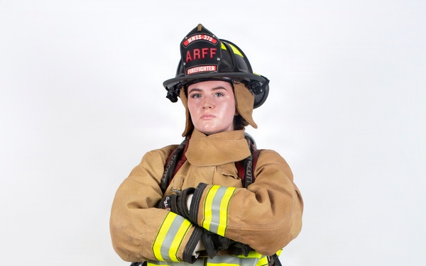 A firefighter in full personal protective equipment (PPE) stands in a confident pose with arms crossed against a plain white background. The individual is wearing tan/gold turnout gear with high-visibility yellow-green reflective striping, a black firefighting helmet labeled 'ARFF' and 'FIREFIGHTER' with unit designation 'MWSS-379'. The image appears to be a professional portrait or official documentation photograph.
