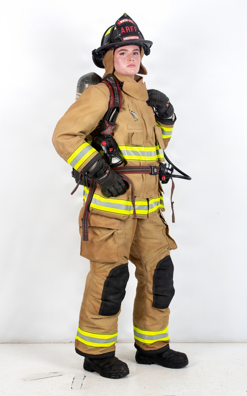 A firefighter in full turnout gear is posed against a white studio background. The individual is wearing complete personal protective equipment including a black helmet labeled 'ARFF' and 'FIREFIGHTER', a tan/gold bunker suit with high-visibility yellow reflective striping, and is carrying an SCBA (Self-Contained Breathing Apparatus) on their back. The image appears to be a professional portrait or equipment showcase photograph.