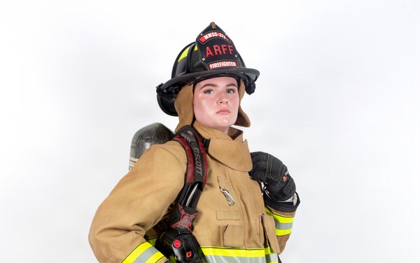 A firefighter in full turnout gear is posed against a white studio background. The individual is wearing complete personal protective equipment including a black helmet labeled 'ARFF' and 'FIREFIGHTER', a tan/gold bunker suit with high-visibility yellow reflective striping, and is carrying an SCBA (Self-Contained Breathing Apparatus) on their back. The image appears to be a professional portrait or equipment showcase photograph.