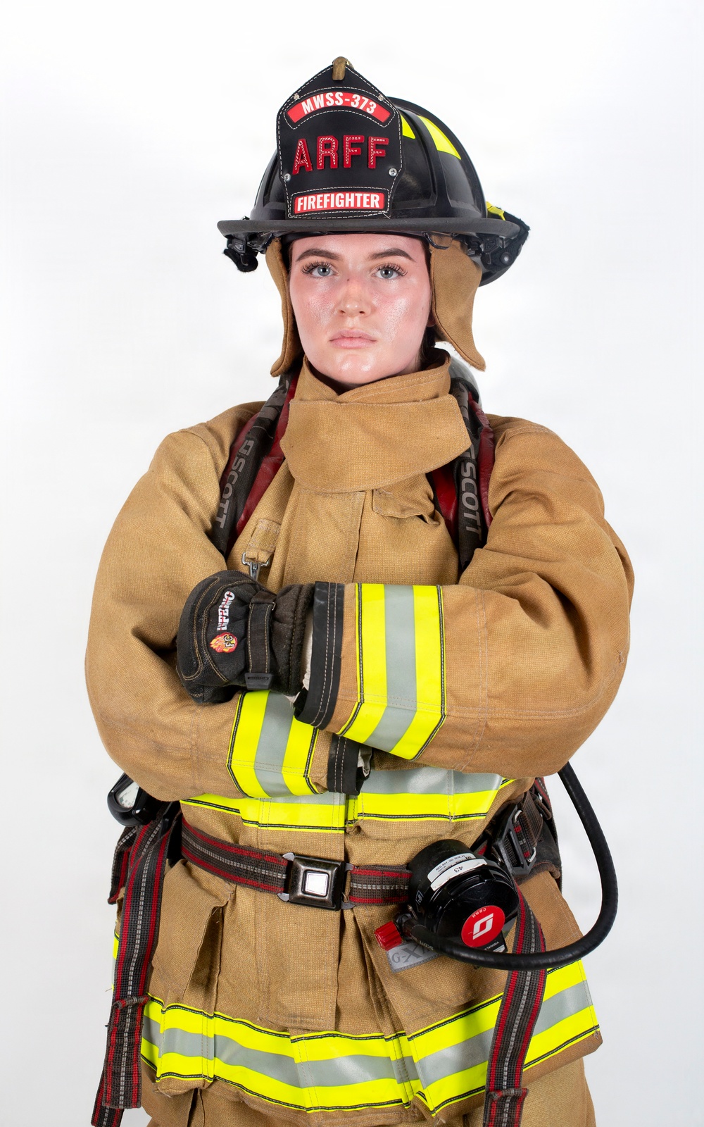 A firefighter in full turnout gear poses against a white background with arms crossed in a confident stance. The subject is wearing a black helmet labeled 'ARFF' and 'MWSS-373' with 'FIREFIGHTER' designation, along with tan/gold structural firefighting gear equipped with SCBA (Scott Air-Pak) harness. This appears to be a professional portrait photograph.