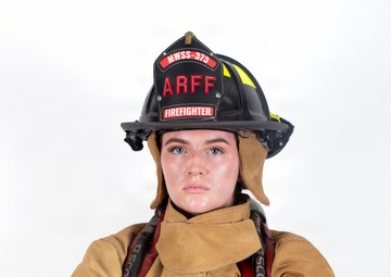A firefighter in full turnout gear poses against a white background with arms crossed in a confident stance. The subject is wearing a black helmet labeled 'ARFF' and 'MWSS-373' with 'FIREFIGHTER' designation, along with tan/gold structural firefighting gear equipped with SCBA (Scott Air-Pak) harness. This appears to be a professional portrait photograph.