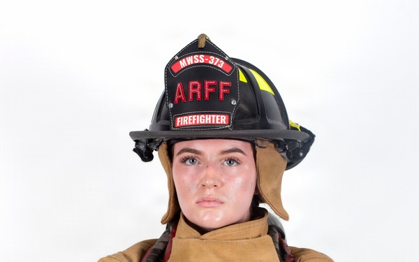 A firefighter in full turnout gear poses against a white background with arms crossed in a confident stance. The subject is wearing a black helmet labeled 'ARFF' and 'MWSS-373' with 'FIREFIGHTER' designation, along with tan/gold structural firefighting gear equipped with SCBA (Scott Air-Pak) harness. This appears to be a professional portrait photograph.