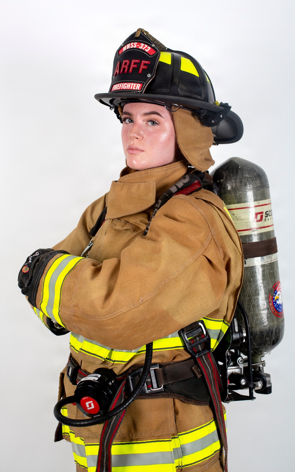 A firefighter in full turnout gear poses for a professional portrait against a white background. The individual is wearing an ARFF (Aircraft Rescue and Firefighting) helmet marked 'MWSS-373' and 'FIREFIGHTER,' with arms crossed in a confident stance. Full personal protective equipment is visible including a SCBA (Self-Contained Breathing Apparatus) tank on the back.