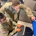 Two U.S. Army soldiers in OCP (Operational Camouflage Pattern) uniforms are working together at a computer workstation. One soldier is seated at the keyboard while the other leans over and points at the monitor screen, appearing to provide instruction or guidance. The setting appears to be a military office or training facility with cubicle-style workstations.