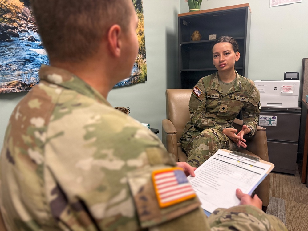 Two U.S. Army soldiers in OCP (Operational Camouflage Pattern) uniforms are engaged in what appears to be a counseling or administrative session in an office setting. One soldier, facing away from the camera, holds a clipboard with a form and appears to be conducting an interview or assessment. The other soldier, a female, is seated in a chair facing the interviewer in what looks like a behavioral health or administrative office.