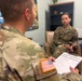 Two U.S. Army soldiers in OCP (Operational Camouflage Pattern) uniforms are engaged in what appears to be a counseling or administrative session in an office setting. One soldier, facing away from the camera, holds a clipboard with a form and appears to be conducting an interview or assessment. The other soldier, a female, is seated in a chair facing the interviewer in what looks like a behavioral health or administrative office.