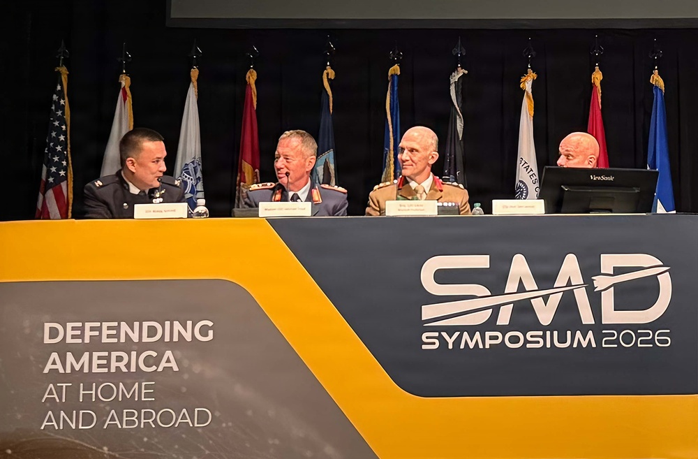 Four military officers are seated at a panel table at the SMD Symposium 2026, an event themed 'Defending America at Home and Abroad.' The panelists appear to be from multiple nations based on their distinct uniforms, suggesting an allied or multinational defense forum. The setting includes multiple military flags displayed behind the panel table.