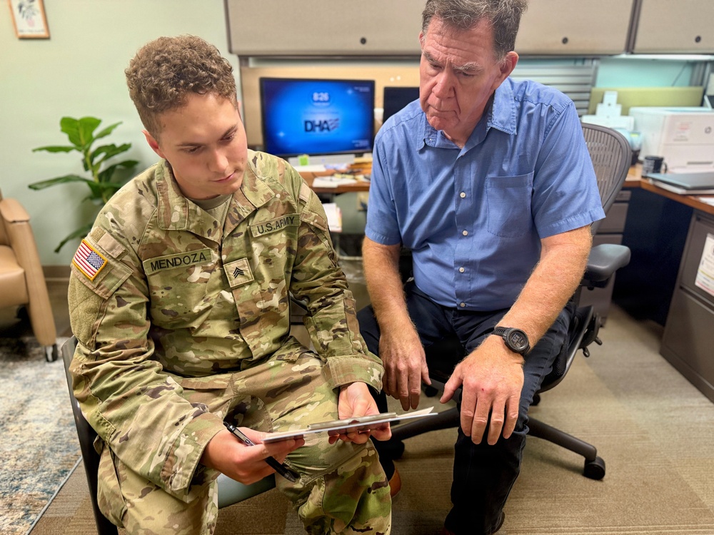 A U.S. Army soldier with the name tape 'Mendoza' and rank of Sergeant is seated in an office environment reviewing a document on a clipboard with a civilian man in a blue shirt. The two individuals appear to be engaged in a consultation or administrative meeting. The setting appears to be a military-affiliated office, with a monitor displaying 'DHA' (Defense Health Agency) visible in the background.