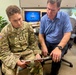 A U.S. Army soldier with the name tape 'Mendoza' and rank of Sergeant is seated in an office environment reviewing a document on a clipboard with a civilian man in a blue shirt. The two individuals appear to be engaged in a consultation or administrative meeting. The setting appears to be a military-affiliated office, with a monitor displaying 'DHA' (Defense Health Agency) visible in the background.