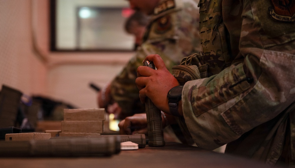 Military personnel in MultiCam/OCP camouflage uniforms are seated at a table handling equipment and materials. The foreground soldier is holding what appears to be a spray can or similar cylindrical object, while another soldier in the background appears to be writing. Various items including what appear to be cardboard boxes and cylindrical objects are laid out on the table.