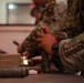 Military personnel in MultiCam/OCP camouflage uniforms are seated at a table handling equipment and materials. The foreground soldier is holding what appears to be a spray can or similar cylindrical object, while another soldier in the background appears to be writing. Various items including what appear to be cardboard boxes and cylindrical objects are laid out on the table.