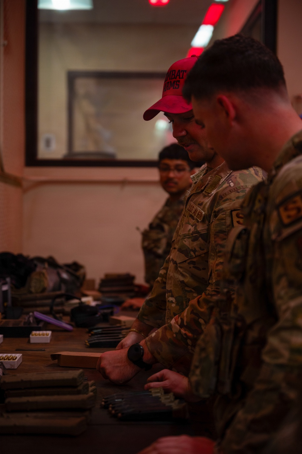 Several military personnel in camouflage uniforms are gathered around a table covered with what appear to be magazines, ammunition boxes, and other weapons-related equipment. One individual is wearing a red cap with text reading 'COMBAT ARMS.' The setting appears to be an indoor armory or weapons issue facility with dim, reddish lighting.