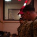 Several military personnel in camouflage uniforms are gathered around a table covered with what appear to be magazines, ammunition boxes, and other weapons-related equipment. One individual is wearing a red cap with text reading 'COMBAT ARMS.' The setting appears to be an indoor armory or weapons issue facility with dim, reddish lighting.