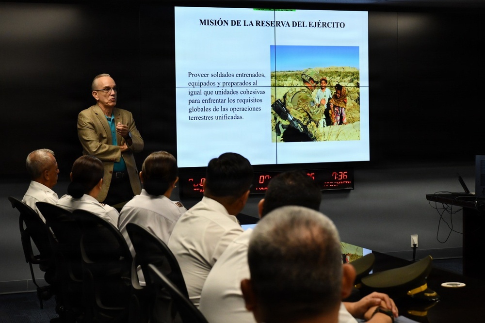 A civilian presenter is delivering a briefing in Spanish to a group of military personnel in a conference or briefing room. The presentation slide displayed on a large screen is titled 'MISIÓN DE LA RESERVA DEL EJÉRCITO' (Mission of the Army Reserve), accompanied by a field photograph of soldiers interacting with civilians. The audience, dressed in white military uniforms, is seated at a curved desk facing the presenter.