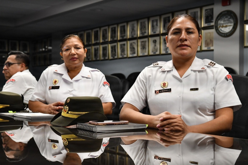 Three uniformed personnel, two women and one man, are seated at a reflective conference table in what appears to be an official military or police meeting room. The foreground woman's name tag reads 'S. HUANASCA C.' and both women wear white uniforms with rank insignia and institutional badges. The room is decorated with framed portraits and a clock on the wall behind them.