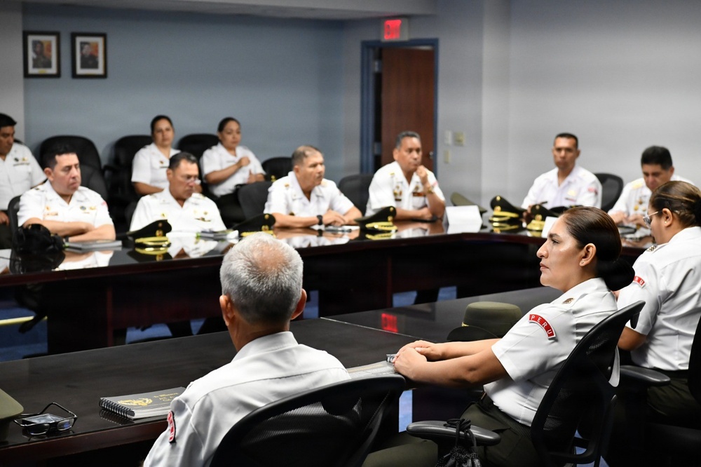 A formal military meeting or briefing is taking place in a conference room, with uniformed personnel seated around a large dark wooden table. The individuals are wearing white military uniforms with visible insignia and patches, including what appears to be 'PERU' country identifiers on some uniforms. Military caps with gold insignia are placed on the table in front of several attendees.