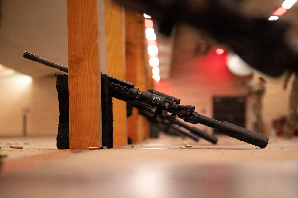 The image shows multiple military-style rifles leaning against wooden dividers on the floor of what appears to be an indoor shooting range. Spent brass casings are visible on the floor near the weapons. In the background, a person in military/tactical clothing can be seen at a firing position.