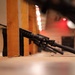 The image shows multiple military-style rifles leaning against wooden dividers on the floor of what appears to be an indoor shooting range. Spent brass casings are visible on the floor near the weapons. In the background, a person in military/tactical clothing can be seen at a firing position.