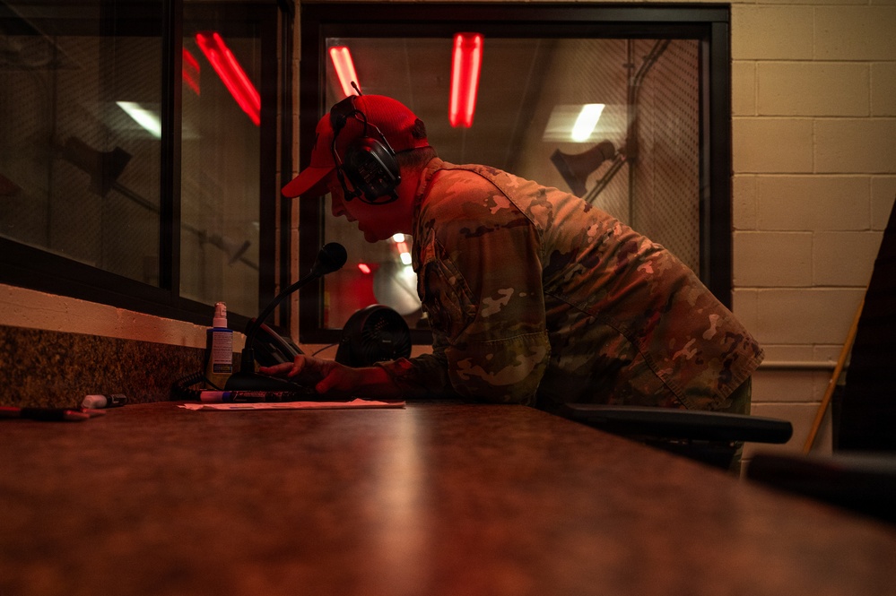 A military personnel member wearing OCP (Operational Camouflage Pattern) uniform and a red baseball cap is leaning over a counter, writing or reviewing documents. The individual is equipped with tactical hearing protection earmuffs and appears to be working at what looks like a range control station or administrative desk inside an indoor shooting range. Red warning lights are visible through a window in the background, consistent with an active firing range environment.