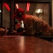A military personnel member wearing OCP (Operational Camouflage Pattern) uniform and a red baseball cap is leaning over a counter, writing or reviewing documents. The individual is equipped with tactical hearing protection earmuffs and appears to be working at what looks like a range control station or administrative desk inside an indoor shooting range. Red warning lights are visible through a window in the background, consistent with an active firing range environment.