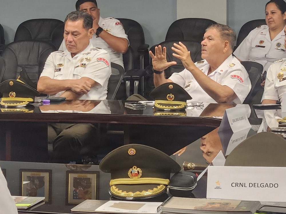 A formal military meeting or conference is taking place, featuring uniformed officers seated at a conference table. The officers are wearing white military dress uniforms with insignia and patches, with their service caps placed on the table. Name placards, including 'CRNL DELGADO' and 'GRAL BRIG VILLASS' (partially visible), along with an 'Army Reserve' logo placard, are visible on the table.